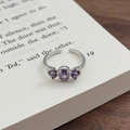 Nahaufnahme eines silbernen Blumenrings mit lila Zirkonia auf einer Buchseite - Einstellbar Offen / Oval Birnenform (JZ1002A)