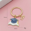 Détail du porte-clés de remise des diplômes 2025 avec bonnet bleu et livre sur fond rose - Combinaison Toque De Diplômé Bleue Et Livres (2025)