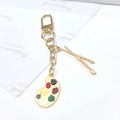 Detalle de un llavero artístico dorado con paleta de pintura y pinceles de arte - Hebilla Con Forma De Puerta De Tablero De Dibujo