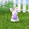 Miniatur-Osterhase-Figur auf Gras sitzend mit einem leuchtend lila Osterei in den Armen - Hase mit lila Ei (Modell C)