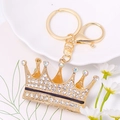 Detalle de un llavero de corona dorada con diamantes de imitación en bandeja blanca - Negro