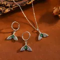 Detailaufnahme von Delfinschwanz-Schmuckset mit grünen Steinen auf braunem Sand. - Fischschwanz mit Einzelnem Grünem Strass / Halsketten- und Ohrring-Set / Silber