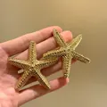 Vista dettagliata di orecchini a stella marina dorati testurizzati tenuti in mano - Forma a stella marina / Oro