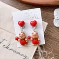 Close-up of cute cartoon bear earrings with red heart and festive party hat on display - Bear Ear Clips