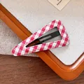 Gros plan d'une barrette BB à carreaux rouges et blancs sur un fond en tissu texturé - Style Triangle