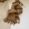 Detail of wool knit scarf in earthy camel tone featuring fringe and a metal logo plaque - Camel