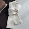 Detail eines cremeweißen Wollstrickschals mit vertikaler Rippenstruktur und Metall-Logo-Tag - Creme