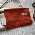 Detailaufnahme der Wollschal-Fransen mit Markenetikett auf warmem karamellfarbenem Stoff - 180*30cm / Melange Orange