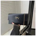 Detail of black envelope clutch featuring a gold bow ornament and fine metal chain strap - Black