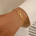 Close-up of a gold-plated infinity symbol bracelet worn on a woman's wrist with a white shirt - Gold Infinity Symbol Bracelet (5975801)