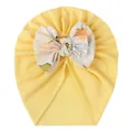 Detailaufnahme einer Babymütze mit großer Blumenschleife auf weichem gelbem Baumwollstoff - Gebrochenes Weiß