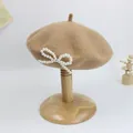 Detail der Wollbaskenmütze in Khaki mit elegantem weißen Perlenbogen auf einem Holzständer - Camel