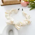 Cream pleated satin headband with elegant bow detail in a soft floral lifestyle setting - Beige