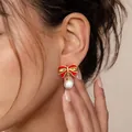 Primer plano de modelo luciendo pendientes de lazo esmaltado rojo y perla blanca - Estilo de lazo de esmalte rojo y perla artificial (e1803) / 14k / Un par / Dorado