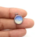 Dettaglio di un ciondolo rotondo argentato con motivo paesaggistico tenuto tra le dita - Opale / 12x15mm