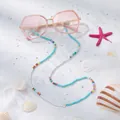 Detail einer Sonnenbrille mit handgefertigter Perlenkette auf sandigem Strandhintergrund - Blau und Weißer Ton