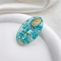 Primer plano de pinza de acetato ovalada con textura de concha verde azulado y logo dorado - Redondo Azul