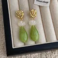Golden rose stud earrings with green waterdrop pendants on a minimalist cream stand - Green