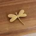Dettaglio della spilla a libellula in oro opaco con rilievi su sfondo in legno scuro - Oro