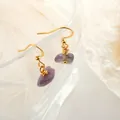 Close-up of handmade gold earrings with raw purple amethyst crystals on a glass edge - Amethyst Gold