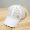 Weiße Baseballkappe aus Spitze mit Blumenmuster und goldenem M-Logo auf Holzhintergrund - Weiß Strass M Logo