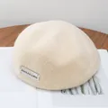 Detailed close-up of cream white wool newsboy hat with minimalist metal logo tag - Adjustable / Beige