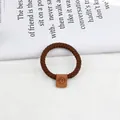 Brown braided hair tie with a small leather smiley face tag on white fabric background - Brown