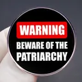 Warning Beware Of The Patriarchy feminist circular metal pin badge held by a gloved hand - Warning Slogan Black