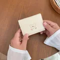 Kurze cremefarbene PU-Ledergeldbörse mit niedlicher Bären- und Blumenstickerei in der Hand - Gebrochenes Weiß