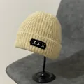 Detail view of oatmeal knit beanie with XXX black fabric patch on a black metal stand - M (56-58cm) / XXX Embroidery Patch Style / Khaki