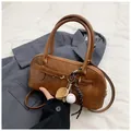 Brown vintage leather shell bag with baseball charm and top handles in a handheld pose - With Pendant / Tan