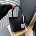 Minimalist black tote bag with light blue heart charm shown on a decorative book background - Black