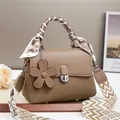 Brown woven crossbody bag with silk scarf handle and floral charm on a display stand - Khaki