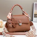 Elegant brown leather handbag with silk scarf handle and floral charm on a white table - Earthy Yellow