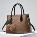 Brown ostrich texture handbag with gold hardware and plush cherry charm on white pedestal - Brown