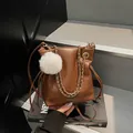 Bolso tipo cubo marrón con cadena dorada, cordón y colgante de peluche sobre libro - Marrón