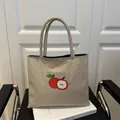 Borsa tote in tela beige con ricamo a mela rossa appoggiata su una sedia moderna - Cachi