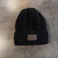 Detail van zwarte kabelgebreide muts met warme oorkleppen en een bruine lederen patch - Zwart