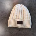 Detail van beige gebreide muts met kabelpatroon en leren patch op de geribde rand - Beige