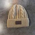 Detail van een beige kabelgebreide muts met een bruine leren patch-accent - Khaki