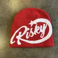 Close-up of red knit beanie with white Risky jacquard lettering on a grey surface - M (54-59cm) / Red