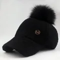 Close-up of black fleece baseball cap with large faux fur pompom and gold M emblem - Adjustable / Black With Black Poms
