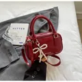Detalle de bolso burdeos brillante con colgante de lazo de perlas sobre chaqueta gris - Burdeos