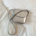 Close-up of a silver metallic crossbody saddle bag with a round buckle on white fabric - Silver