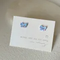 Macro detail of iridescent blue-purple butterfly stud earrings on a white card display - Bow Shape / Lake Blue