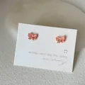 Makroaufnahme kleiner rosa irisierender Schmetterlings-Ohrstecker auf weißer Karte - Schleifenform / Orange