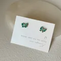 Nahaufnahme der grünen Schmetterlings-Ohrstecker mit goldenem Glitzer auf weißer Karte - Schleifenform / Cyan