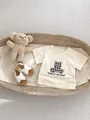 Flat lay of baby beige t-shirt with plaid bear patch and white strappy sandals in a basket - 66CM / Apricot Bear