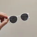 Kids round white frame sunglasses with dark grey lenses held against a neutral wall - White Frame / Black Lens
