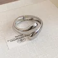 Detalle de un brazalete abierto plateado con forma de gota retorcida en fondo beige - Plateado